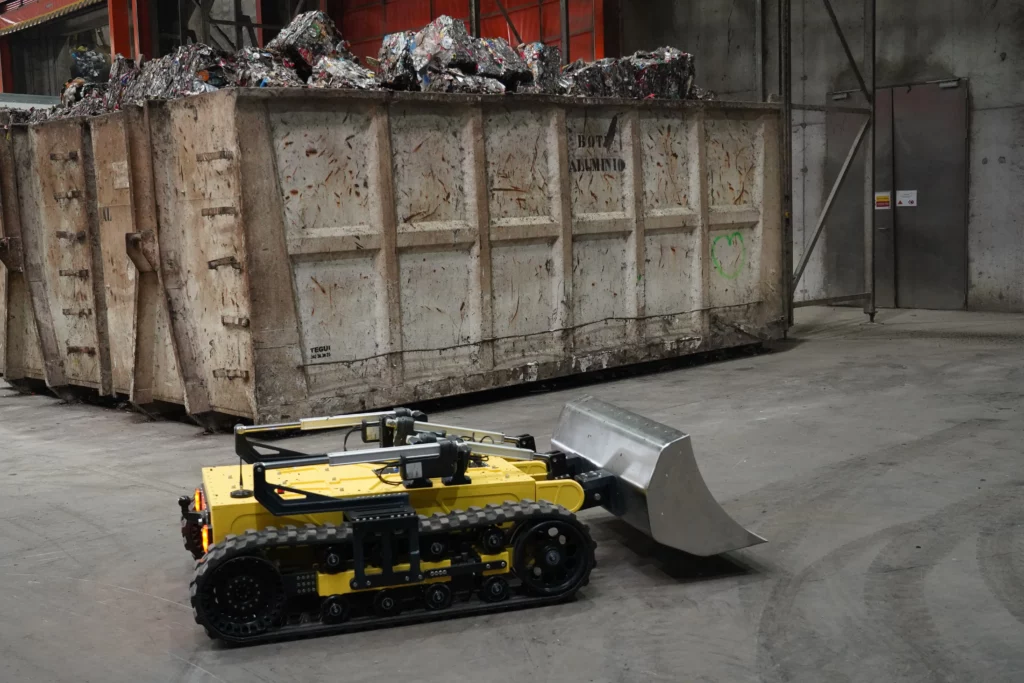 Robot Algor de Sfynx Industry en démonstration dans une déchetterie, équipé d’une lame frontale pour le déplacement et le nettoyage des déchets, évoluant devant une benne de matériaux compressés. L’image illustre l’automatisation des tâches pénibles en centre de tri grâce à un robot chenillé robuste et téléopéré.