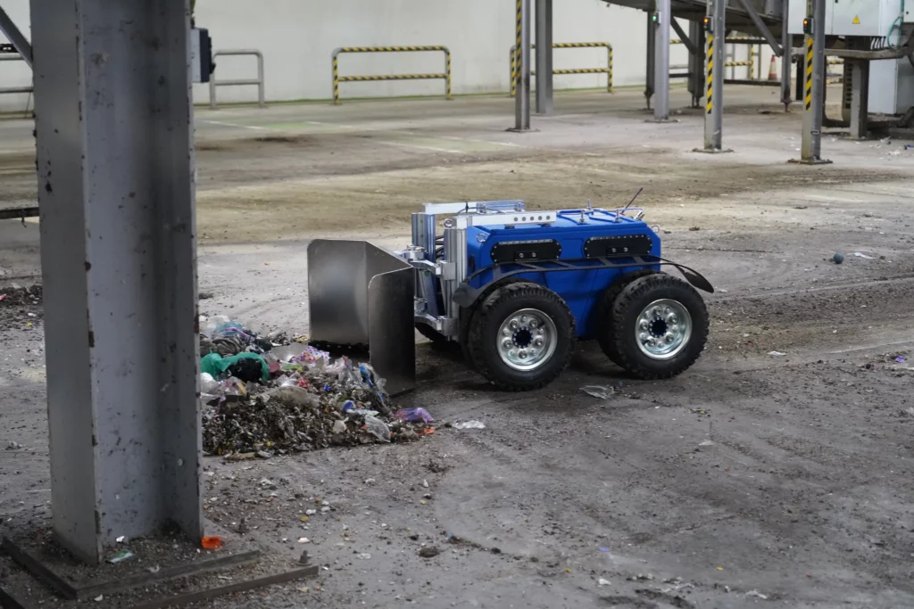 Robot Quator de Sfynx Industry en démonstration dans une déchetterie, utilisant sa lame frontale pour pousser et regrouper des déchets au sol. Le robot tout-terrain à quatre roues illustre l’automatisation des opérations de nettoyage et de manutention dans les centres de tri et environnements industriels.