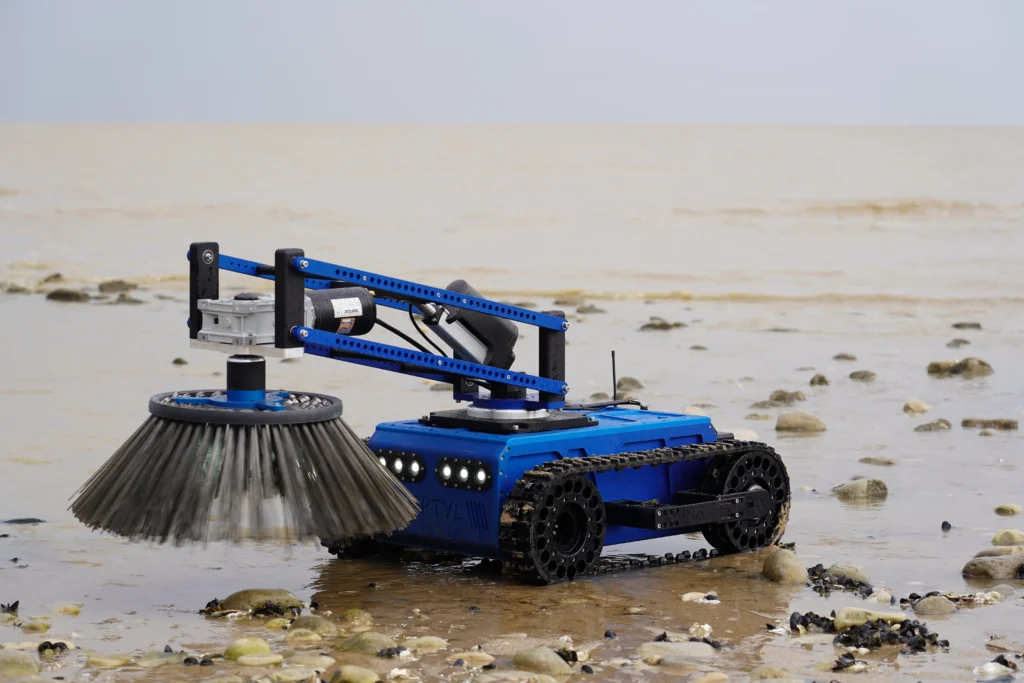 Robot Reptyl V1 de Sfynx Industry équipé d’une brosse rotative, en train de nettoyer une plage en bord de mer sur un terrain humide et rocailleux.