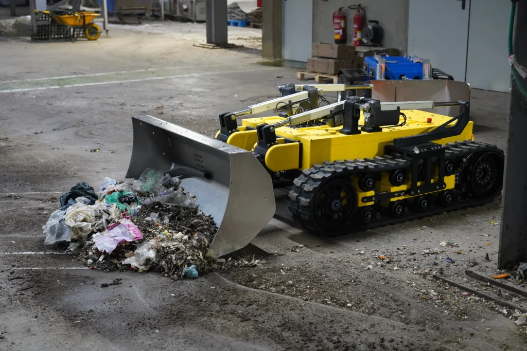 Robot ALGOR de Sfynx Industry en pleine opération de nettoyage, utilisant son large godet frontal pour ramasser des déchets et déblayer le sol dans un environnement industriel.