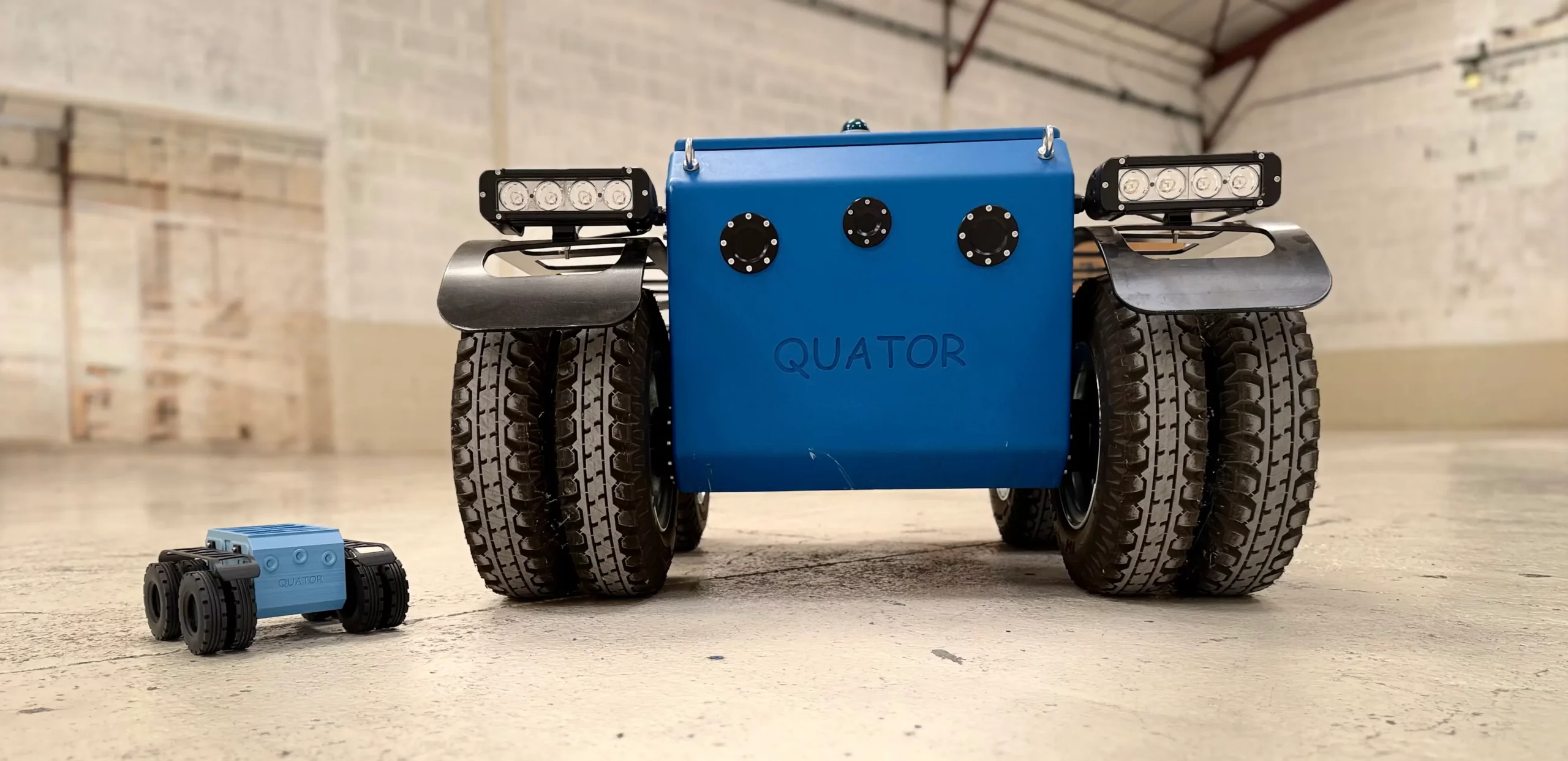 Robot Quator-R de Sfynx Industry présenté en atelier, avec un grand modèle tout-terrain à quatre roues épaisses et un mini-modèle réduit placé à côté, illustrant la modularité, la robustesse et l’échelle comparative de cette plateforme robotique polyvalente.